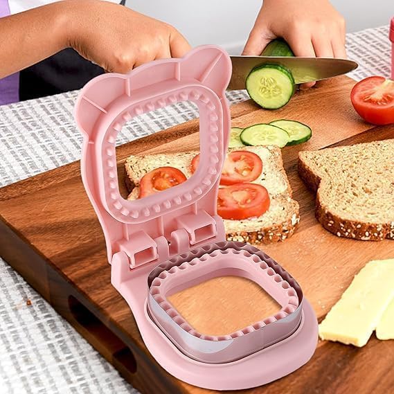 A pink bear-shaped sandwich maker mold is shown open on a wooden cutting board, with bread, sliced tomato, cucumber, and cheese nearby. Two hands are visible holding the mold and slicing vegetables, so this is a Lifestyle image rather than a clean Product/Packaging shot.