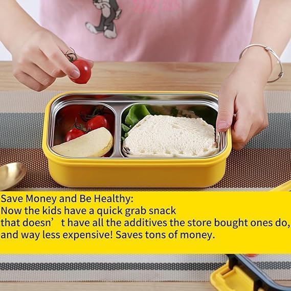 A yellow compartment lunch box with stainless steel trays, shown on a table with a person placing a tomato and holding the lid. The lunch box contains tomato wedges, a slice of cheese, and a sandwich.