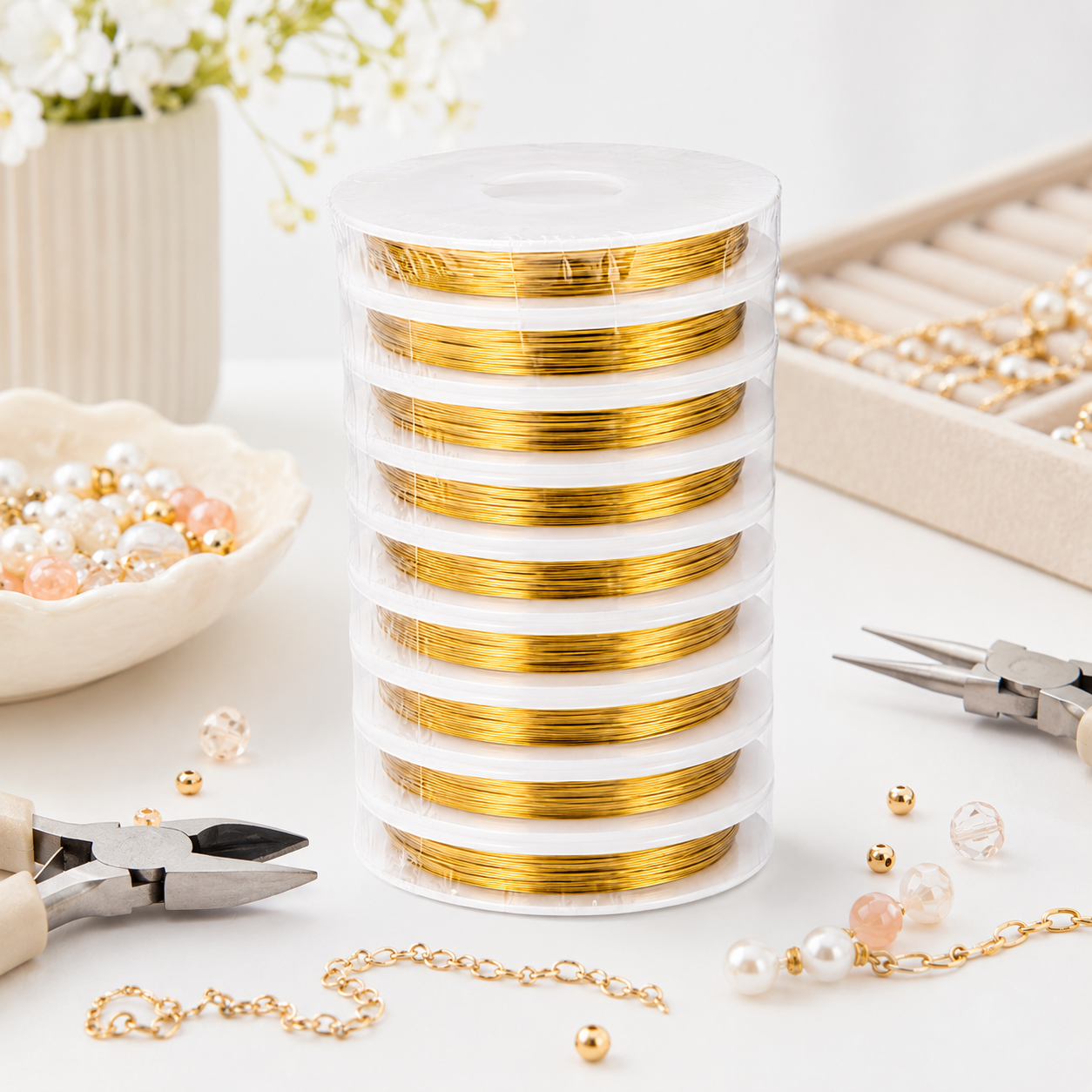 A stack of spools of gold wire wrapped in clear plastic, set on a white surface surrounded by jewelry-making tools, beads, and pearls.