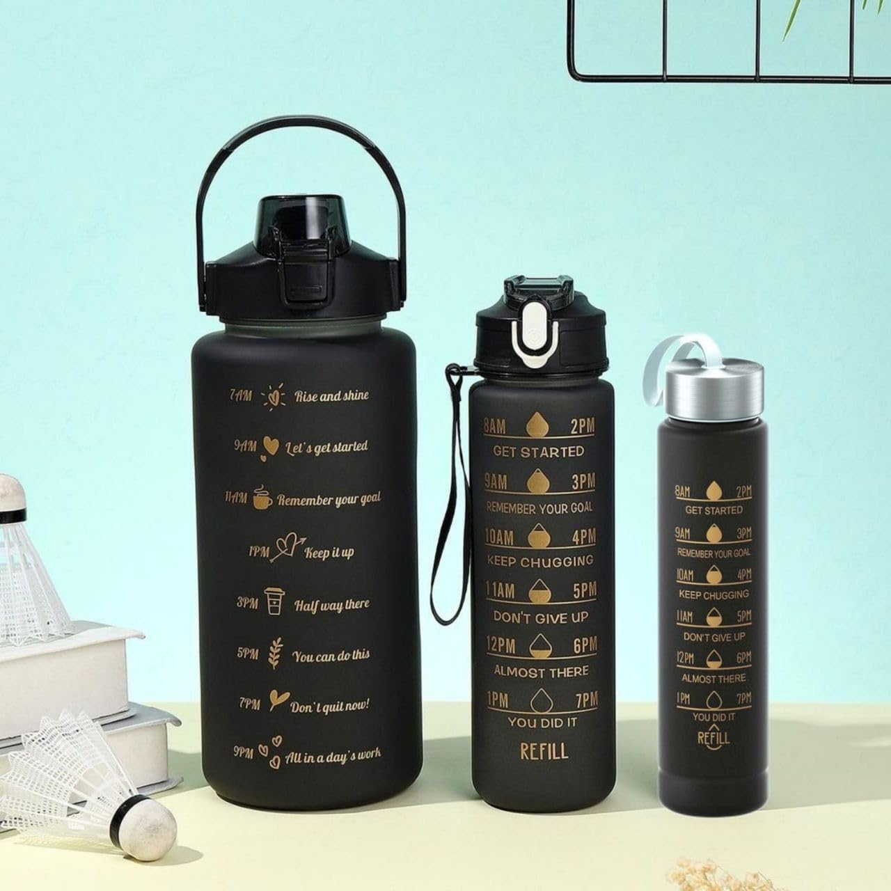 Three black insulated water bottles with gold motivational text and icons, displayed on a light beige surface against a pale blue background.
