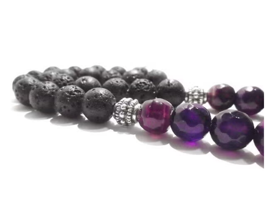 A close-up of a beaded strand with matte black porous lava stone beads transitioning into faceted purple amethyst beads, separated by small silver-toned spacer beads, on a white background.