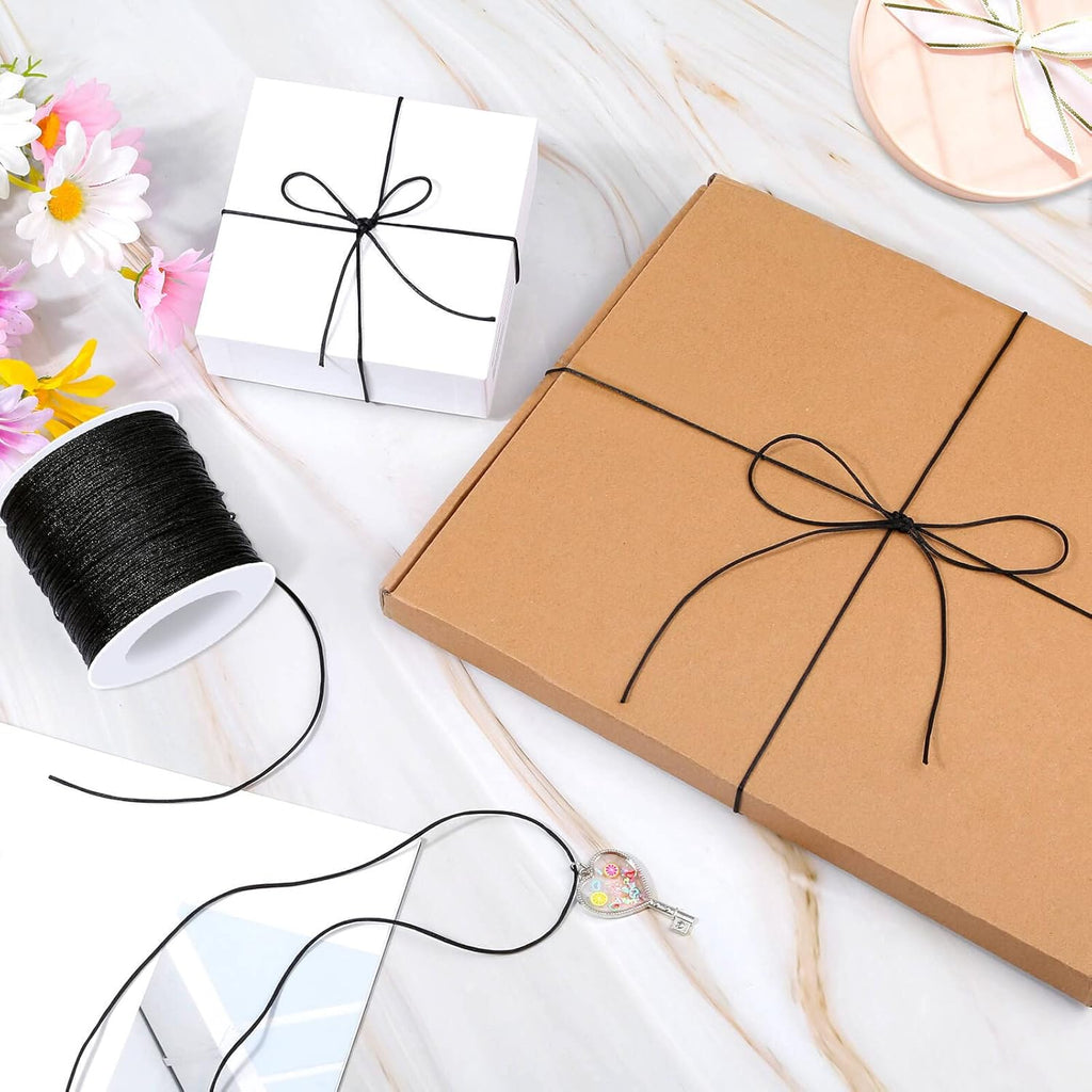 A kraft brown gift box wrapped with black string in a bow, shown on a white marble surface alongside a spool of black thread, a small white gift box, flowers, and a decorative key.