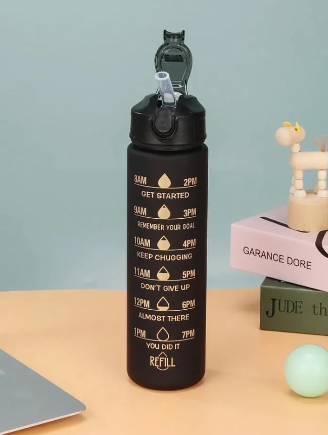 A black reusable water bottle with a flip-top lid and straw, printed with gold time markers and motivational phrases around the circumference. The bottle is shown on a desk next to a laptop edge, books, and a small decorative figurine against a soft blue-green background.