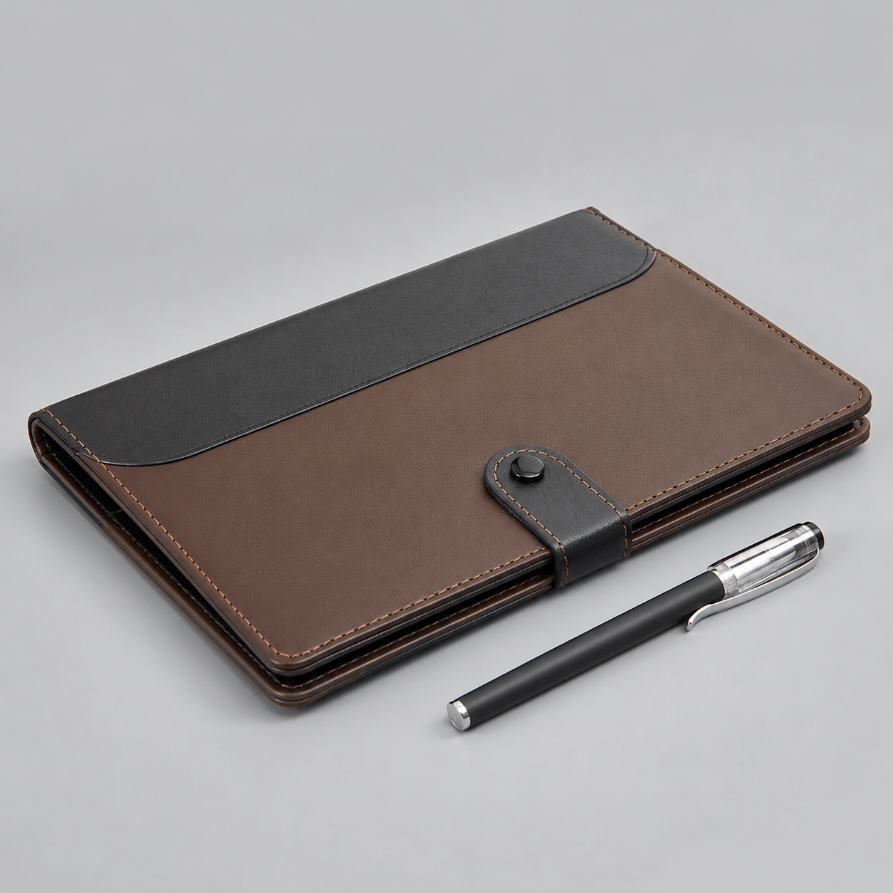 A brown and black leather notebook cover with a pen, photographed on a neutral gray background.