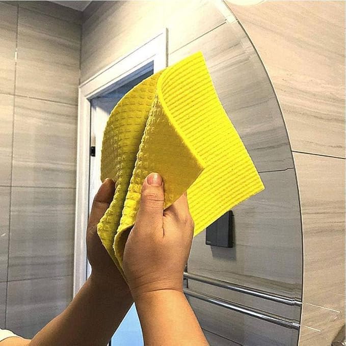A person holding a bright yellow textured cleaning cloth in front of a bathroom mirror with light gray marble-look tiles.