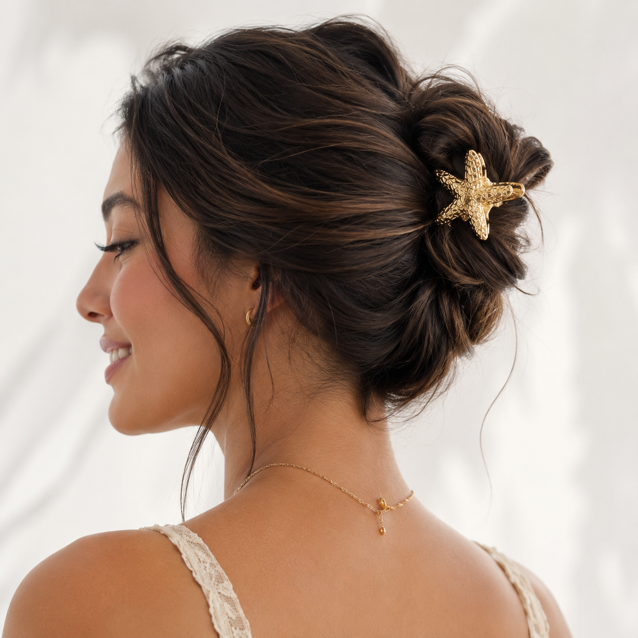 A woman shown from the back and side profile wearing a gold starfish hair claw clip holding her dark brown hair in a low bun, with a delicate gold necklace visible against a soft white background.