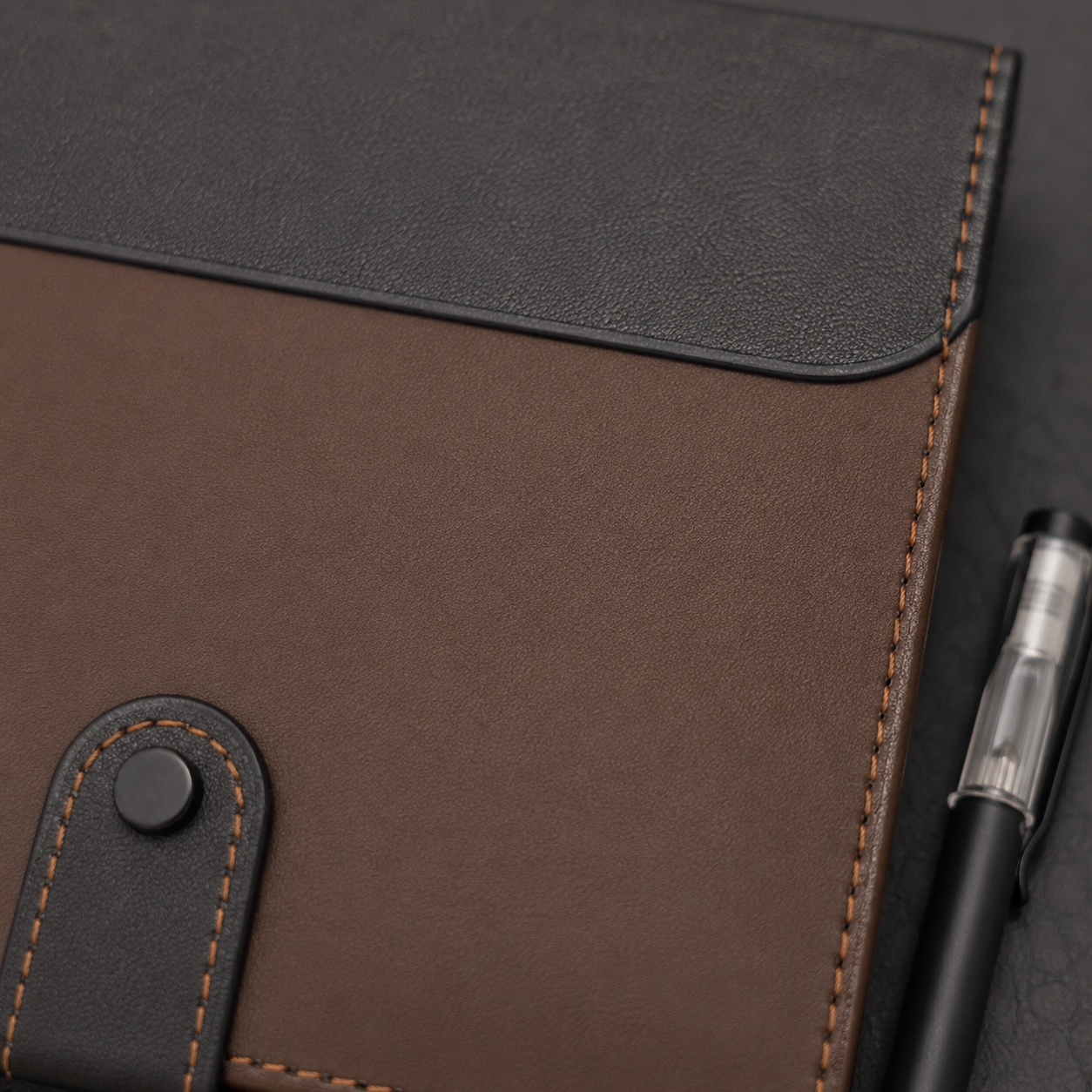 A close-up of a brown and black leather notebook cover or planner sleeve with contrast stitching, a snap-tab closure, and a black pen placed beside it on a dark textured surface.