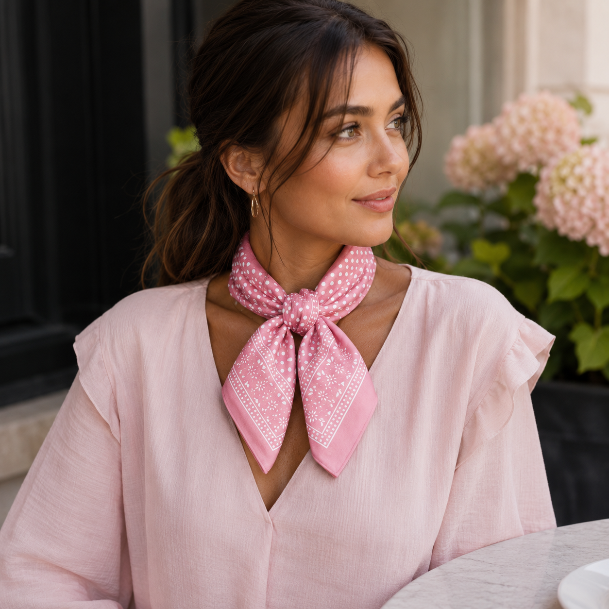 A woman wearing a light pink blouse and a pink patterned neck scarf tied around her collar, posed in front of a soft-focus background with pink flowers.