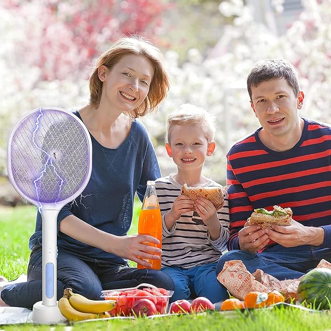 A family of three sits on a grassy lawn outdoors, eating sandwiches and drinking an orange beverage. A purple electric mosquito repellent fan is displayed prominently in the foreground on the left.