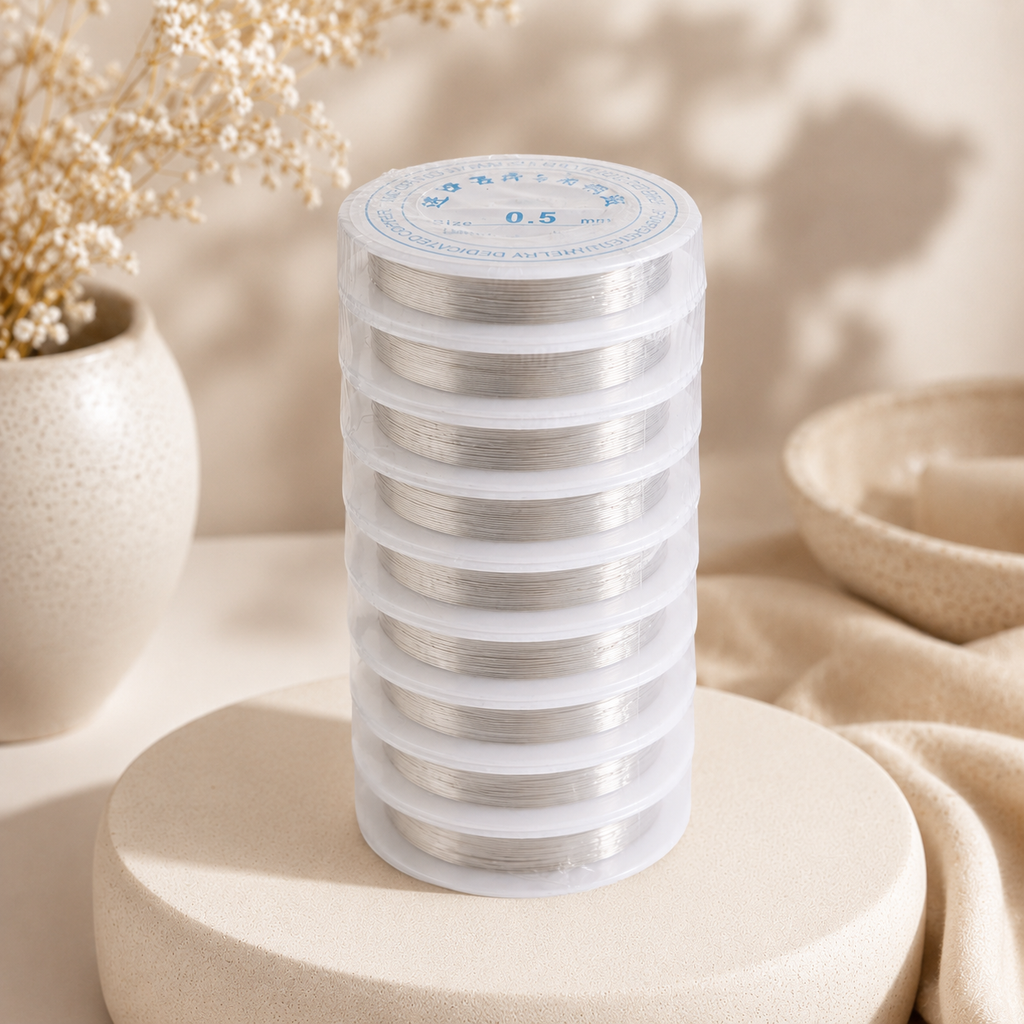 A stack of clear plastic spools containing silver metallic thread, displayed on a round beige pedestal with dried flowers and neutral fabric in the background.