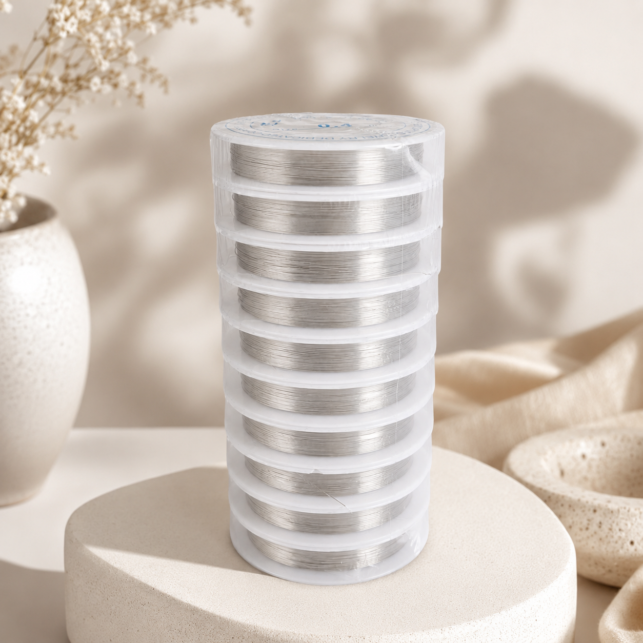 A stack of clear plastic-wrapped spools of silver metallic thread or wire stands upright on a round beige pedestal. The setting is a styled studio scene with soft shadows, a ceramic vase with dried flowers, and neutral fabric in the background.