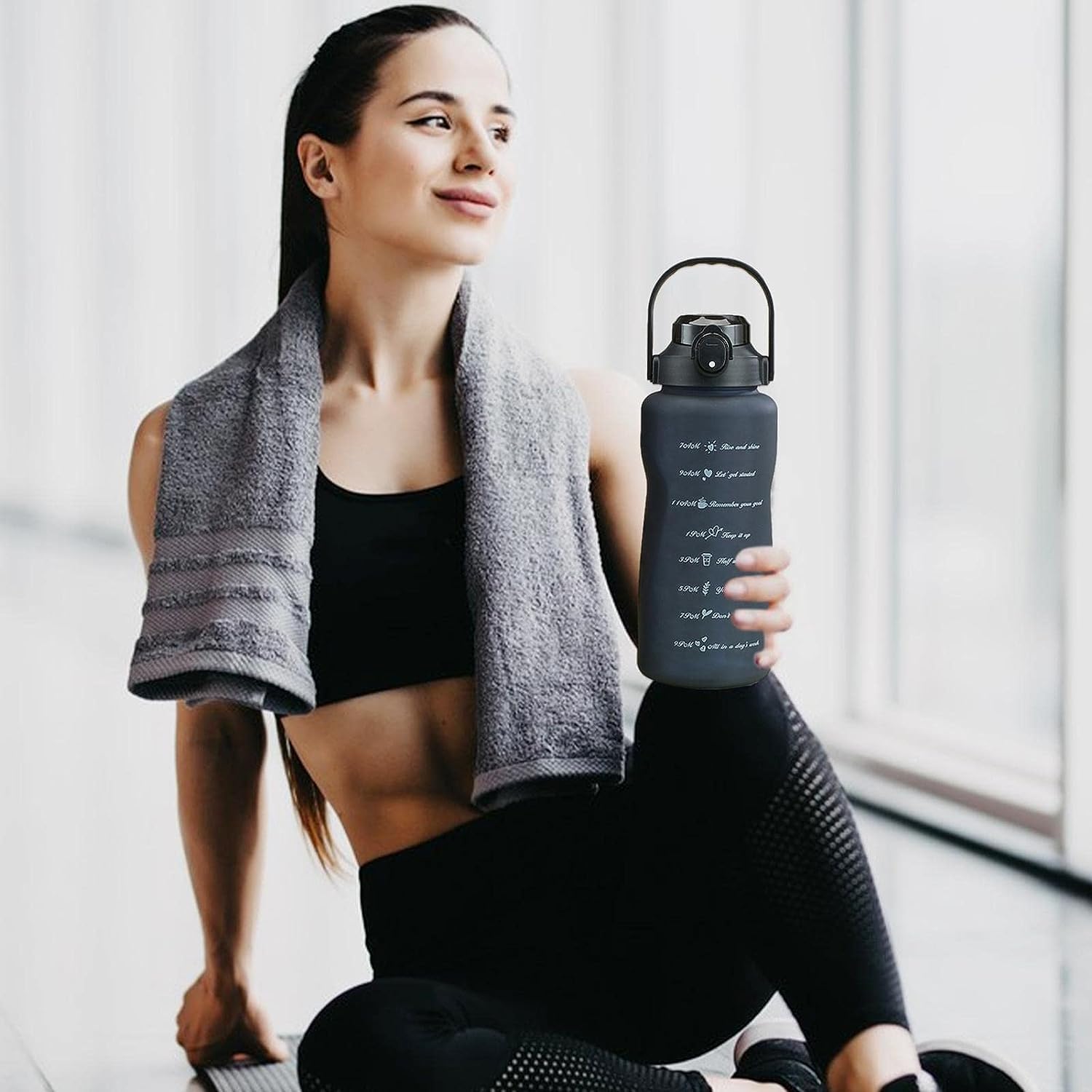 A woman in black athletic wear sits indoors near a bright window, holding a dark gray reusable water bottle with a black handle. A gray towel is draped over her shoulders.