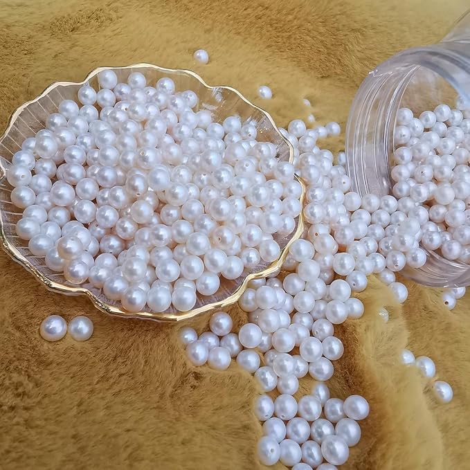 White faux pearl beads with a glossy finish, shown spilling from a clear plastic jar and a gold-rimmed scalloped dish onto a soft tan textured background.
