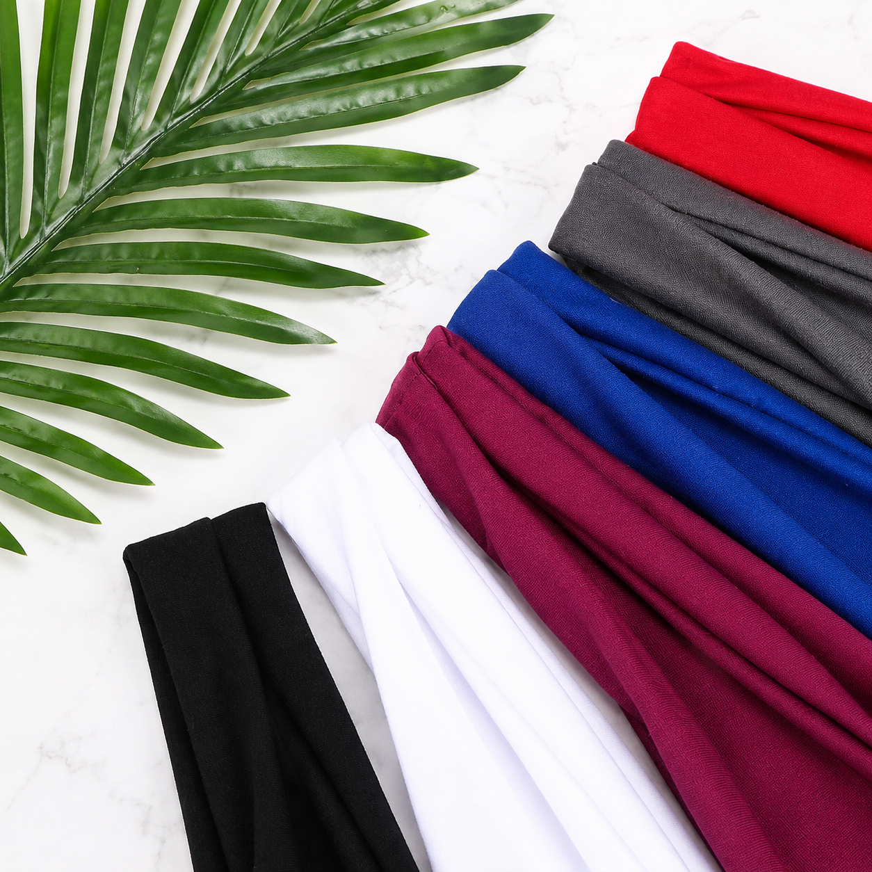 Colorful fabric headbands بألوان red, gray, blue, burgundy, white, و black مرتبة بشكل diagonal بجانب tropical palm leaf على خلفية white marble، بأسلوب stylish flat lay مناسب لعرض fashion & accessories products.
